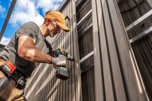 A worker in a cap and gloves uses a power drill to securely fasten lamella panels to a framework under a bright blue sky.