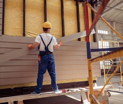 Back view of young man builder wearing safety helmet and work overalls while holding wooden plank and installing exterior wood siding