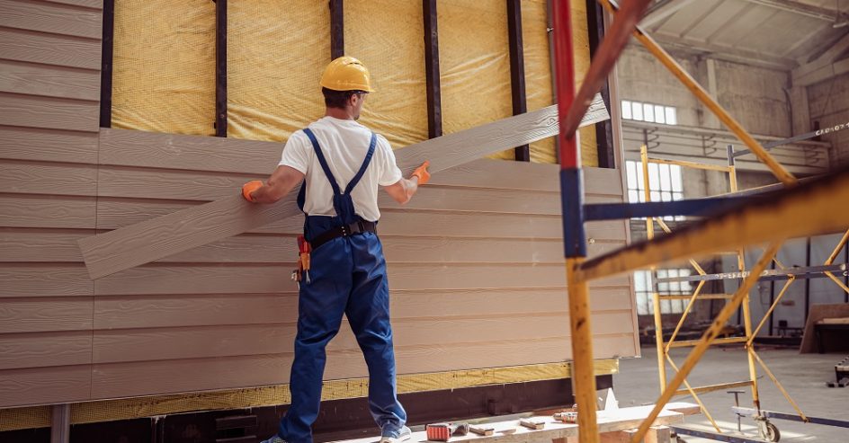 Back view of young man builder wearing safety helmet and work overalls while holding wooden plank and installing exterior wood siding